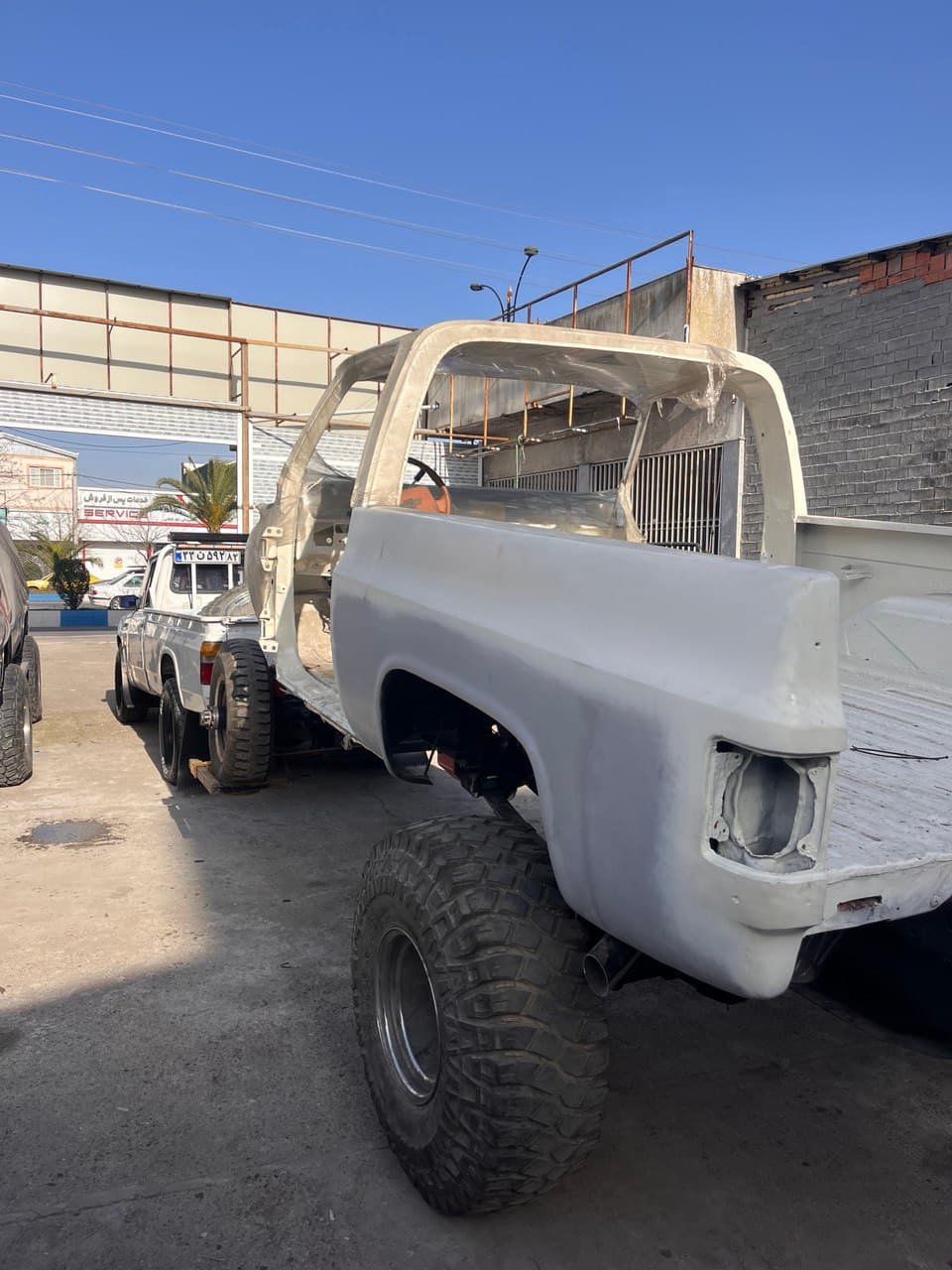 Stripped white truck body with massive off-road tires being towed through an industrial lot.