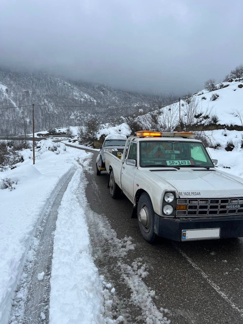White Nissan tow truck pulling a vehicle on a narrow, snowy mountain road.