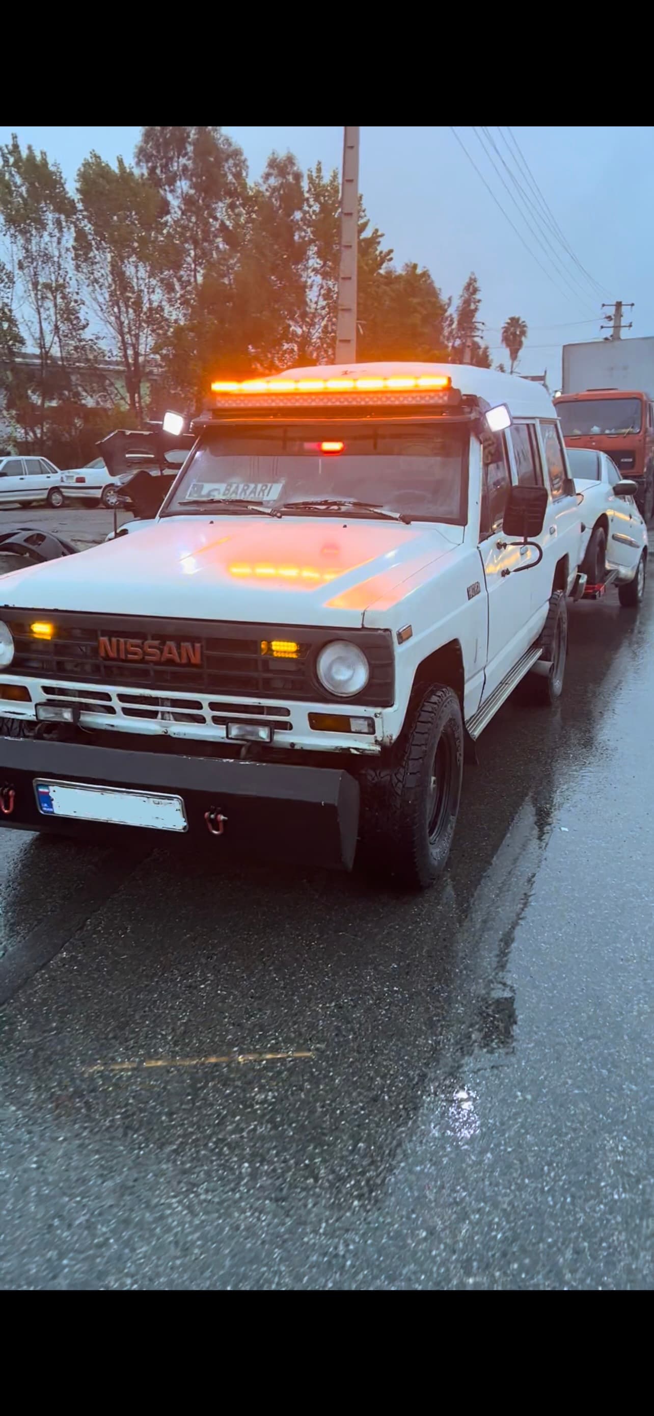 White Nissan Patrol tow truck with amber light bars towing a car on wet pavement.